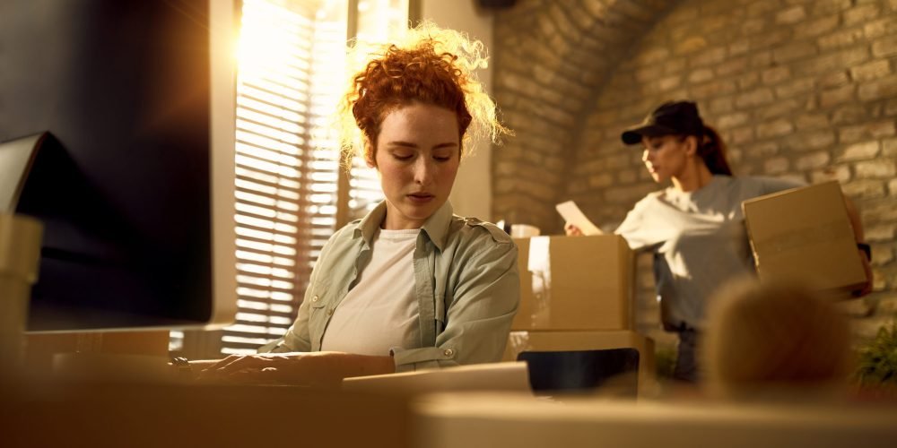 Female courier using computer while working with delivery packag Young woman updating order details while preparing packages for a delivery in the office.